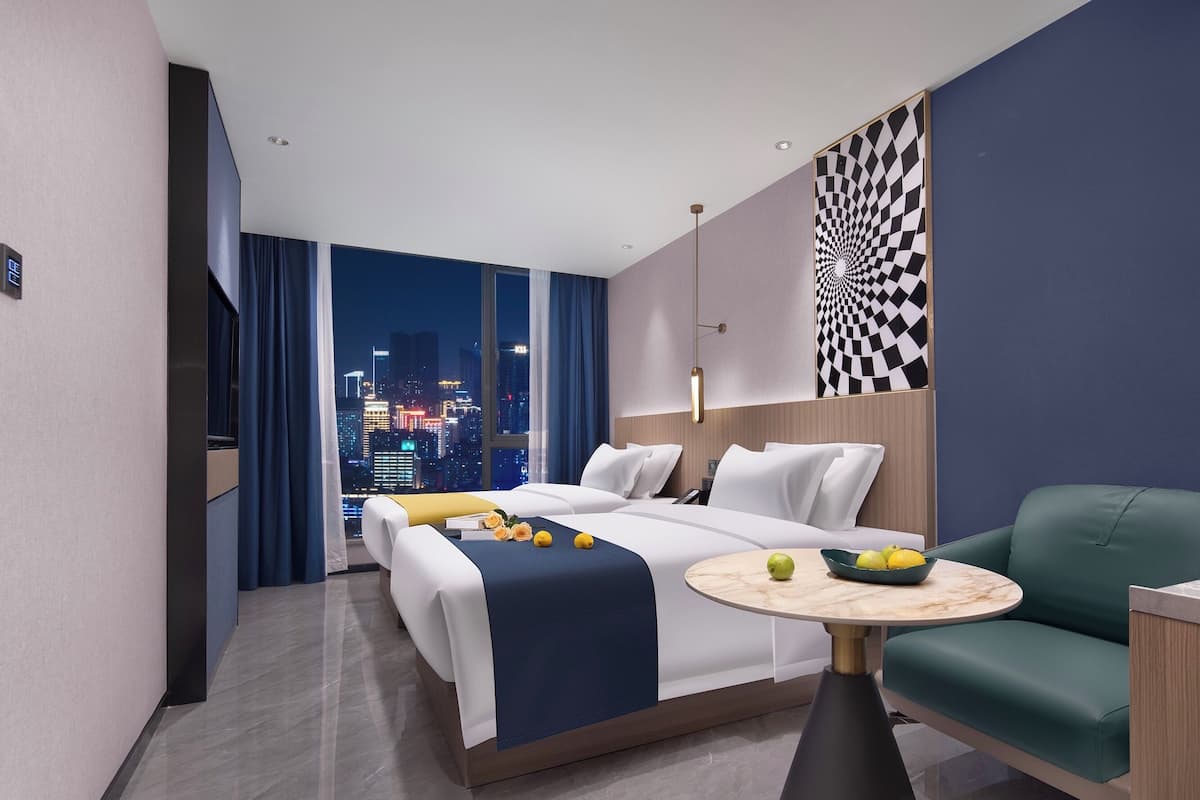 Quarto de hotel moderno com uma cama grande, um sofá verde, uma mesa redonda pequena e vista para a cidade à noite.