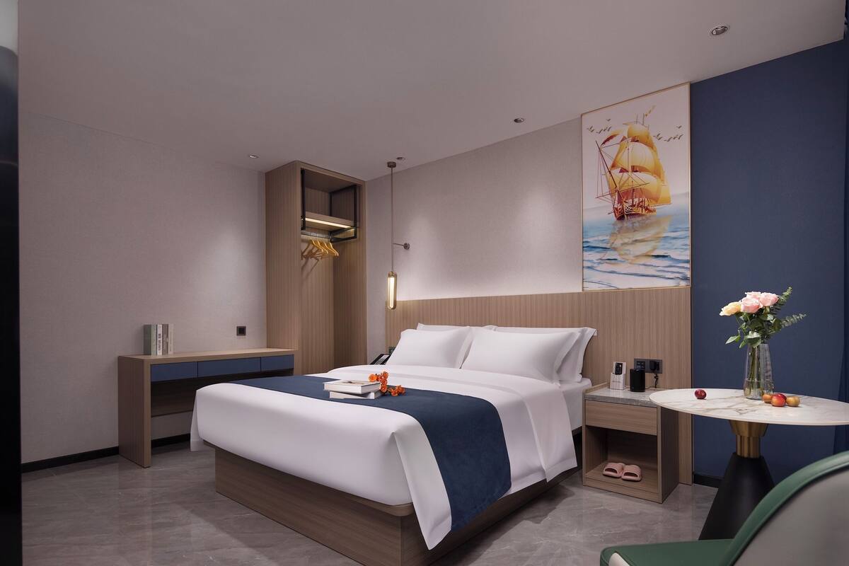 Quarto de hotel moderno com uma cama grande, mesa de cabeceira e uma pequena mesa redonda com um vaso de flores.