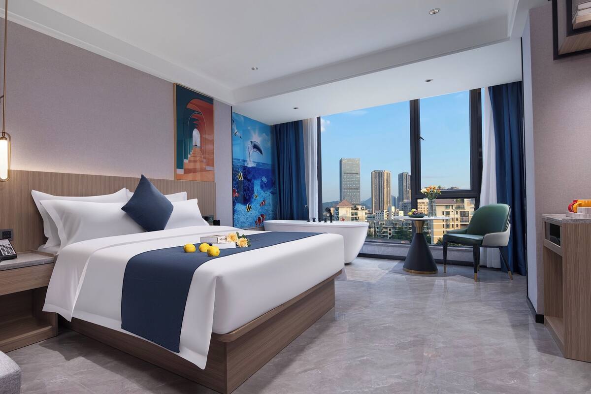 Quarto de hotel moderno com uma cama grande, área de estar e vista para o horizonte da cidade.