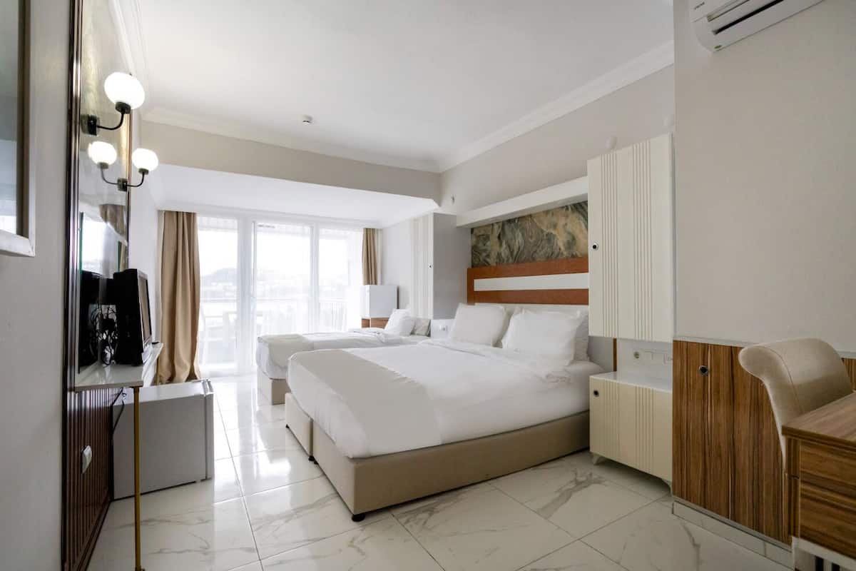 A hotel room with a large bed, a desk with a chair, a wardrobe, and a view of the city through large windows.