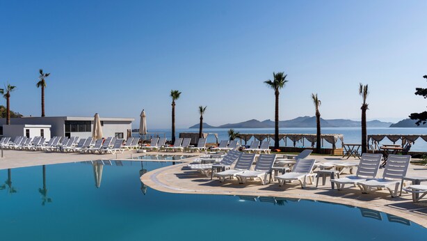 A pool with lounge chairs and palm trees, a building with a white facade, and a clear view of the sea and mountains in the background. - Veluna Beach Resort Yalikavak (Bodrum)