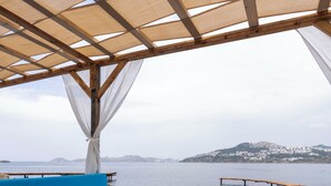 An outdoor dining area with a wooden table and blue cushioned seating, overlooking a body of water and a distant hill. - Veluna Beach Resort Yalikavak (Bodrum)