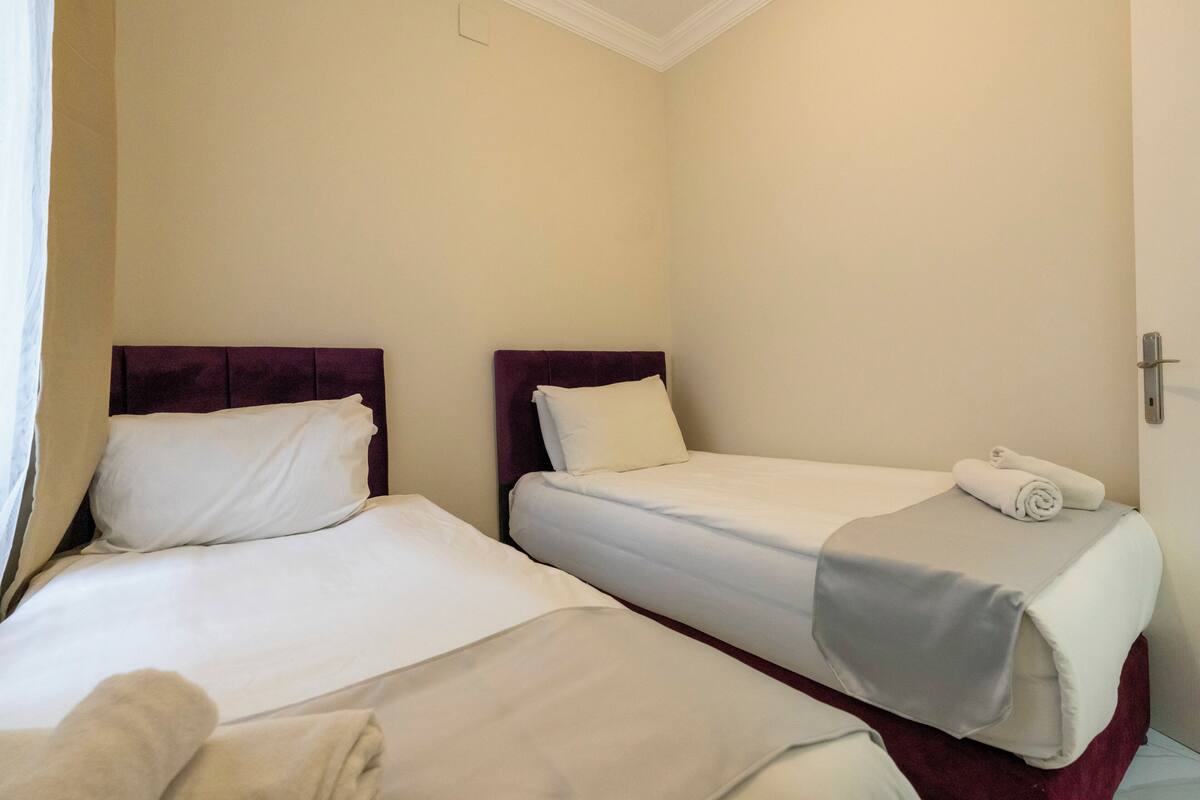 A hotel room with two single beds, each with a headboard, white bedding, and a rolled towel on the bed.
