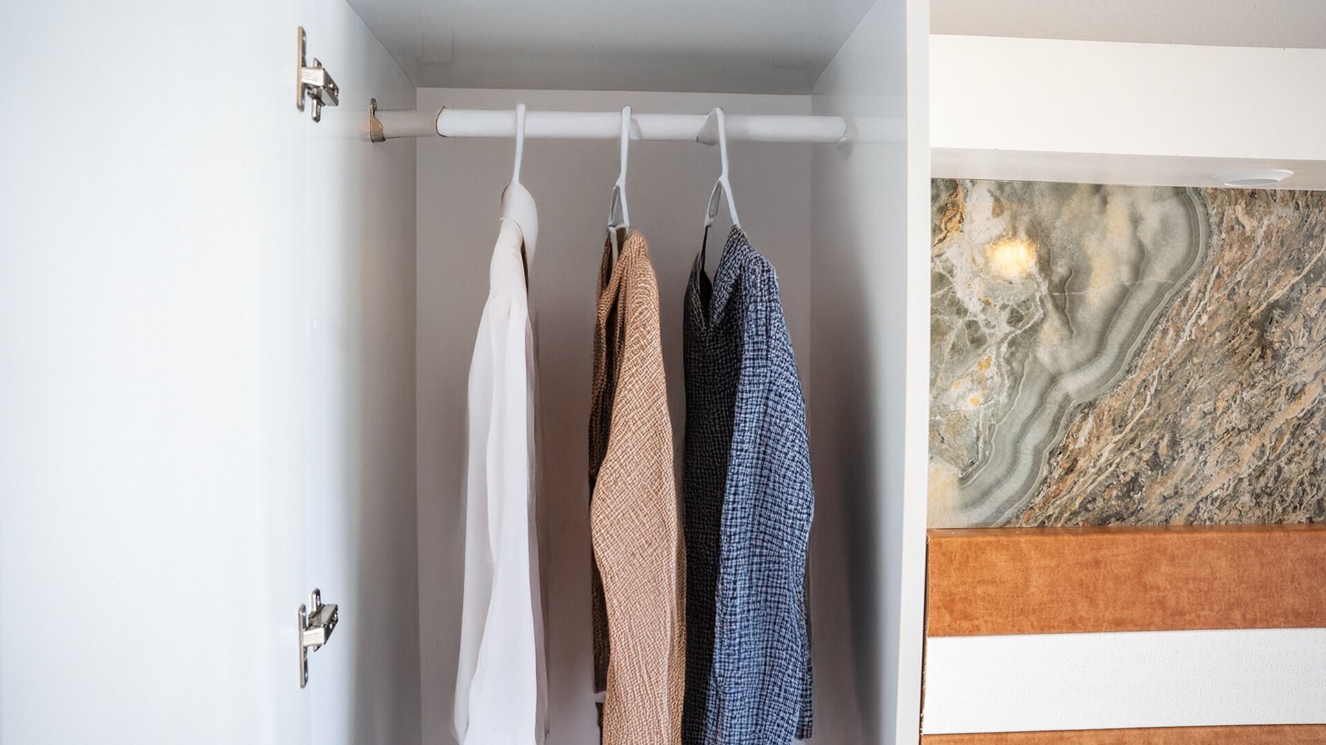 A closet with three hanging garments, a marble wall, and a wooden panel.