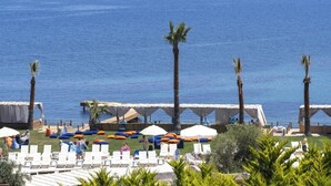 A coastal resort with lounge chairs and umbrellas, a palm tree-lined beach, and a view of the sea and distant buildings. - Veluna Beach Resort Yalikavak (Bodrum)