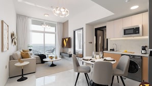 Ein modernes Wohnzimmer mit einer Couch, einem Couchtisch und einem Essbereich. Es gibt eine Küche mit einer Mikrowelle und einer Waschmaschine. Der Raum hat große Fenster und bietet einen Blick auf die Stadt.