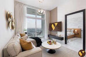 Une chambre d’hôtel moderne dotée d’une grande fenêtre, d’un canapé, d’une table basse ronde sur laquelle sont disposés des fruits, d’une télévision et d’un balcon avec vue.
