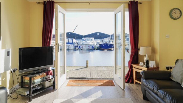 A room with a TV, a sofa, a lamp, and a view of boats at a marina. - Swift Cottage, Norfolk Broads riverside holiday rental with boat mooring (Potter Heigham)