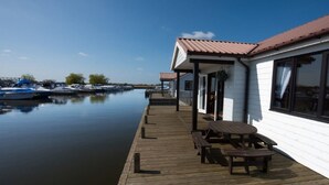 A waterfront property with a wooden deck, a covered seating area, and a building with large windows. - Swift Cottage, Norfolk Broads riverside holiday rental with boat mooring (Potter Heigham)