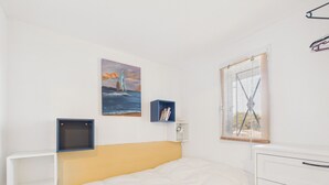 A bedroom with a bed, a bedside table, a nightstand, a window with blinds, a wall-mounted shelf with books, and a painting of a sailboat on the wall. - Sea view, beach 300 m away, apartment for 5 (Saint-Hilaire-de-Riez)