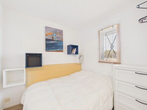 Une chambre à coucher comprenant un lit, une table de chevet, une commode, une fenêtre avec des stores, une étagère murale avec des livres et un tableau représentant un voilier accroché au mur.
