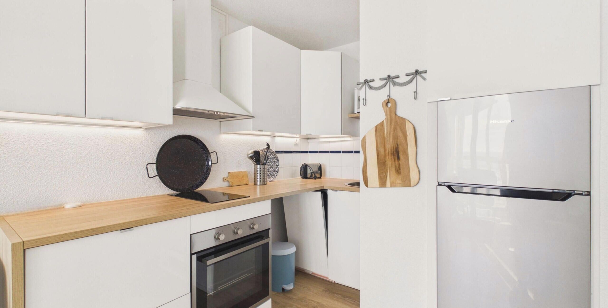 A modern kitchen with white cabinetry, a wooden countertop, a black frying pan, a cutting board, and a stainless steel refrigerator.