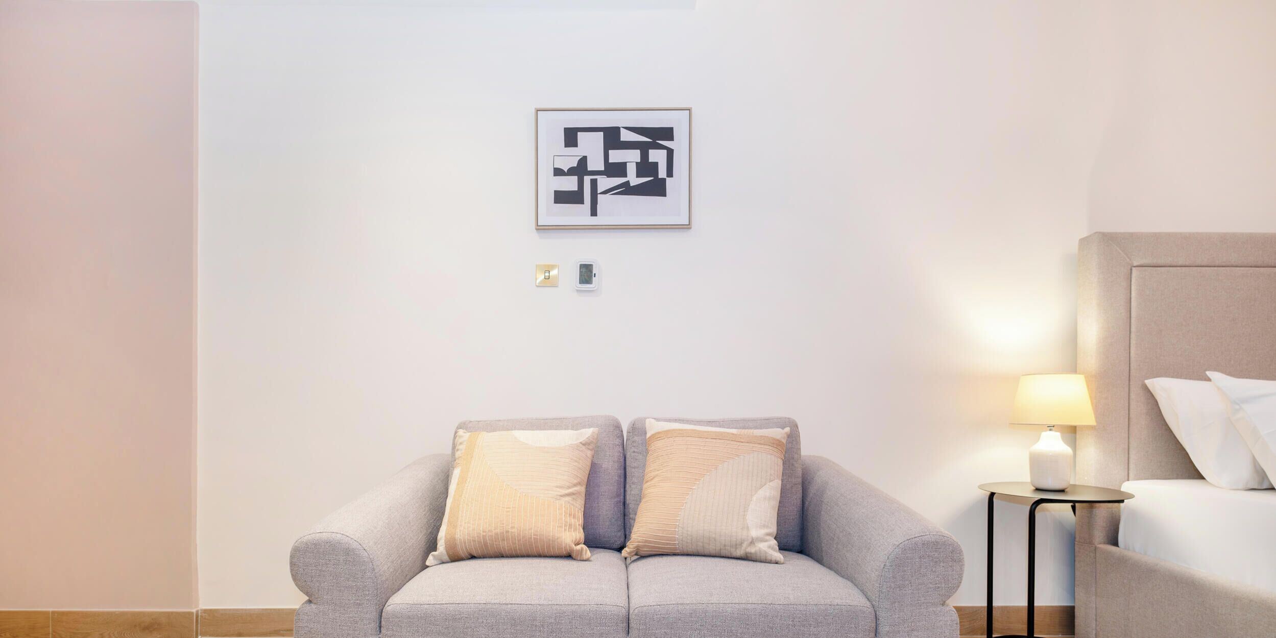 A modern hotel room with a grey sofa, a bedside table with a lamp, and a framed abstract artwork on the wall.