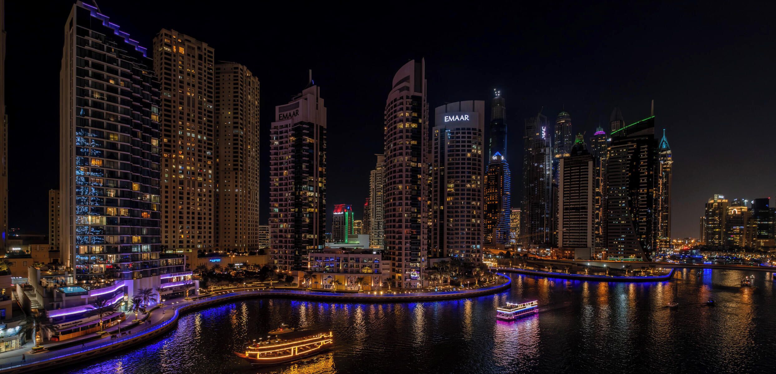 A nighttime cityscape with illuminated high-rise buildings and a river with a boat.