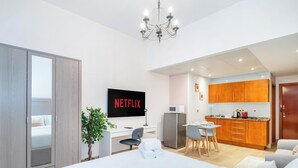 A modern hotel room with a bed, a desk with a computer, a TV displaying 'NETFLIX', a small kitchen area, and a dining table with chairs. - LUX The Marina View Suite 2 (Dubai)