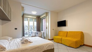 A hotel room with a bed, a yellow sofa, a TV, and a window with a view of the outside. - Bright Studio Apartment with Private Garden (Bergamo)
