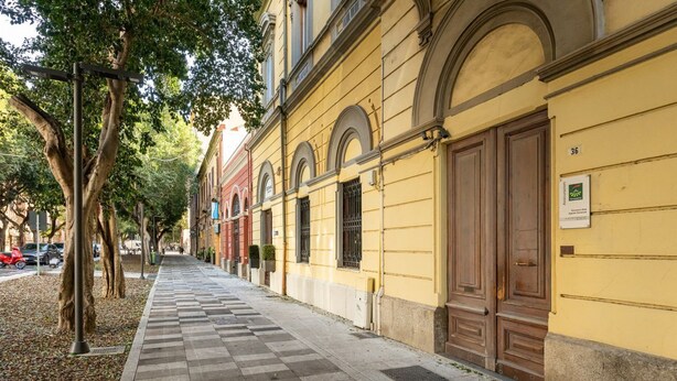 A street with a checkered sidewalk, trees, and a yellow building with large wooden doors. - Trieste Avenue Apartment (Cagliari)