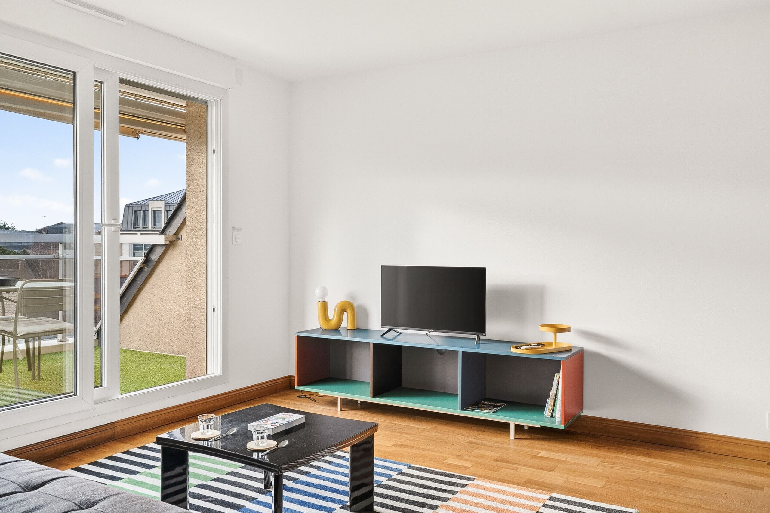 A modern living room with a large sliding glass door leading to a balcony, a flat-screen TV on a teal shelf, and a coffee table with a striped rug.