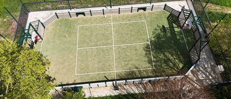 An aerial view of a fenced tennis court with a net, surrounded by greenery and trees.