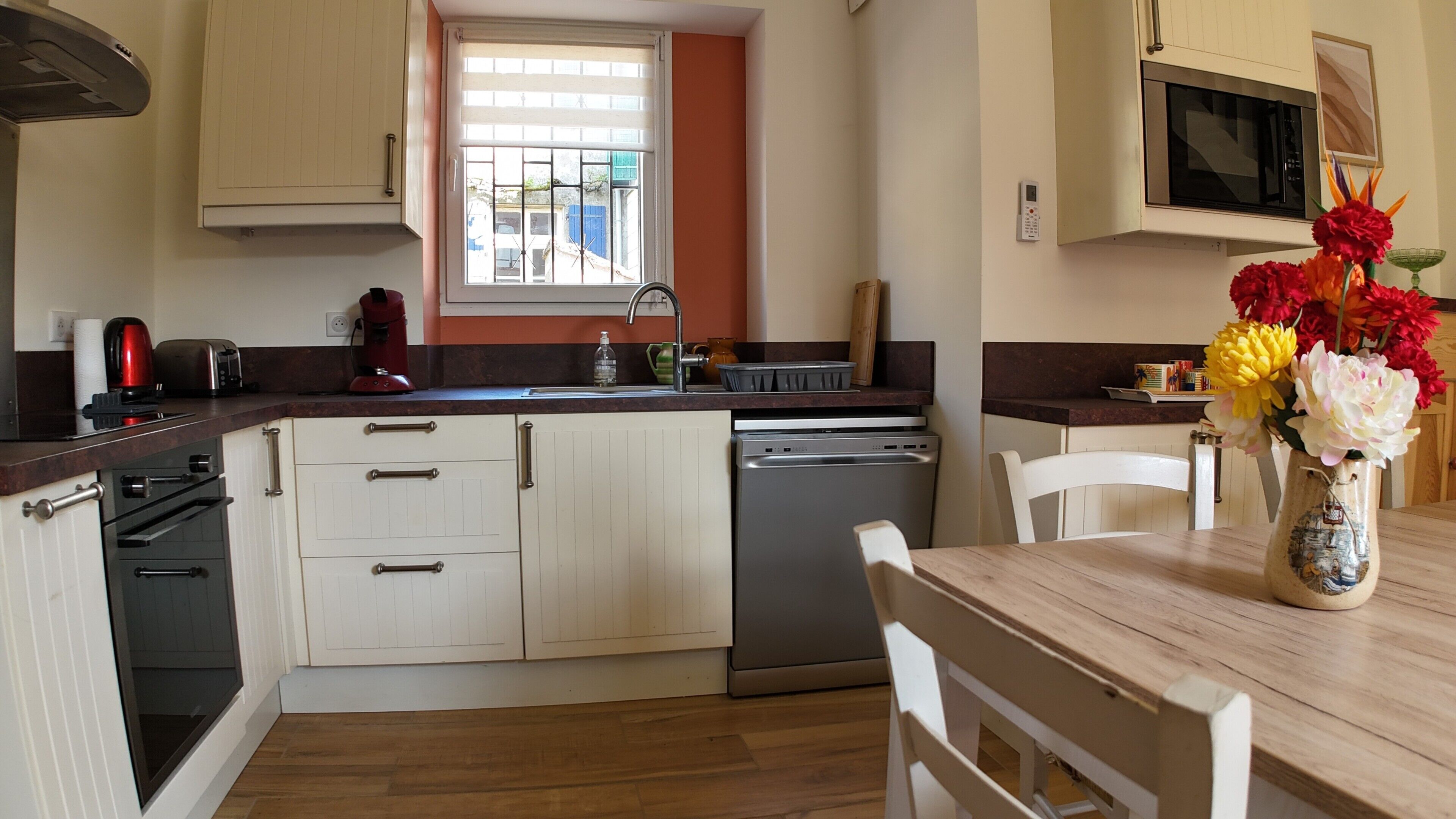 A kitchen with a dishwasher, a toaster, and a coffee maker. There is a window with blinds, a microwave, and a vase with flowers on the table.