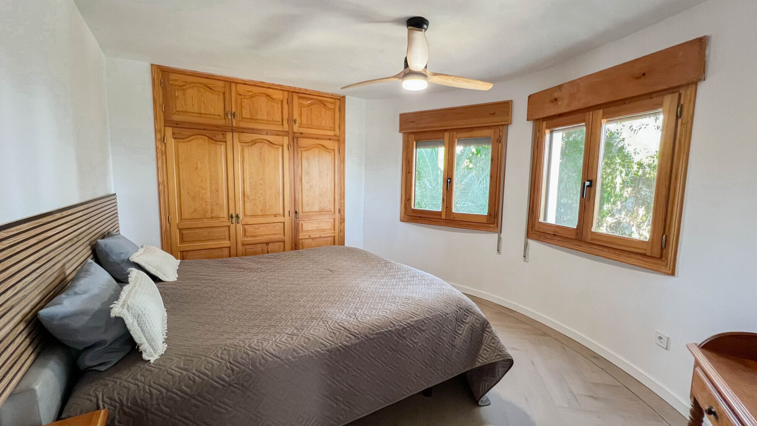 Ein Schlafzimmer mit einem Bett, einem Holzschrank, zwei Fenstern und einem Deckenventilator.