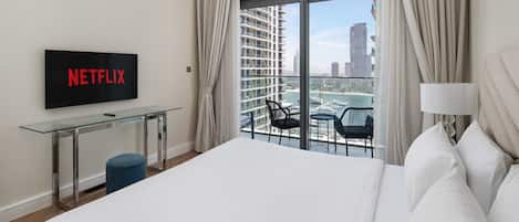 A hotel room with a bed, a TV displaying 'NETFLIX', a glass table, a chair, and a view of a cityscape through large windows.