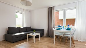 Ein modernes Wohnzimmer mit einem grauen Sofa, einem kleinen weißen Tisch mit Pflanze und einem Essbereich mit weißen Stühlen und blauen Tellern.