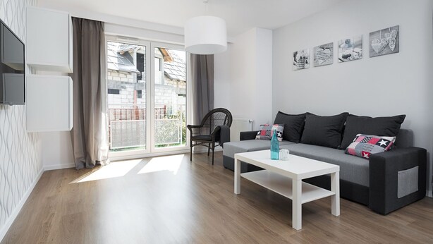 Ein modernes Wohnzimmer mit grauem Sofa, einem weißen Couchtisch und einem schwarzen Sessel. Vier Bilder hängen an der Wand, ein großes Fenster mit Vorhängen und eine weiße Deckenleuchte sind ebenfalls zu sehen.