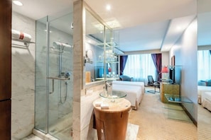 Une chambre d’hôtel moderne dotée d’une douche en verre, d’une coiffeuse ronde en bois avec un lavabo en verre, d’un lit avec une couette blanche et d’un coin salon avec une table en verre.