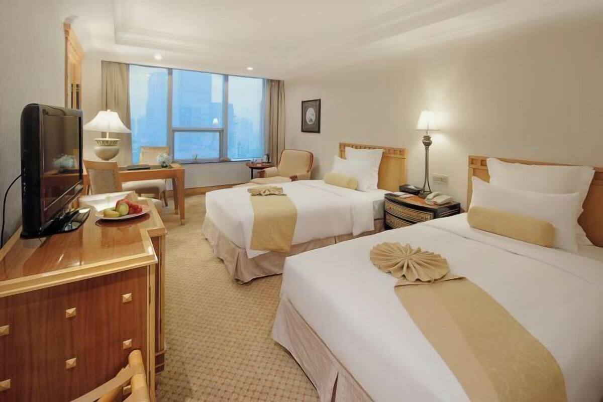 Habitación de hotel con dos camas, un escritorio con televisor, una silla y ventana con vista a la ciudad.