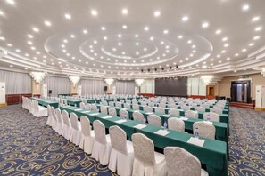 Une salle de conférence avec des rangées de tables et de chaises, un grand écran et un sol recouvert de moquette.