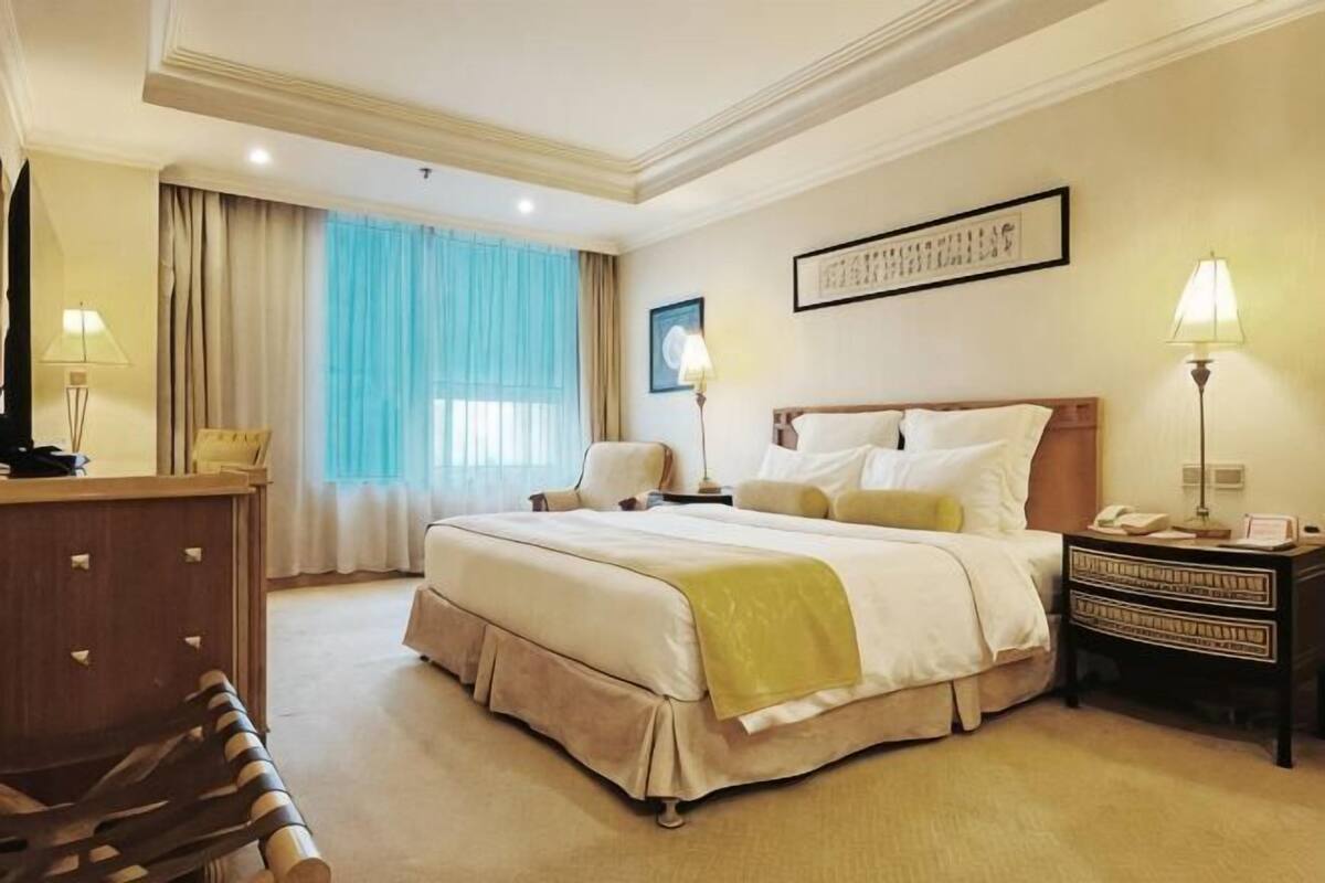 Habitación de hotel con una cama grande, un escritorio, una silla, una mesita de noche y un ventanal con cortinas.