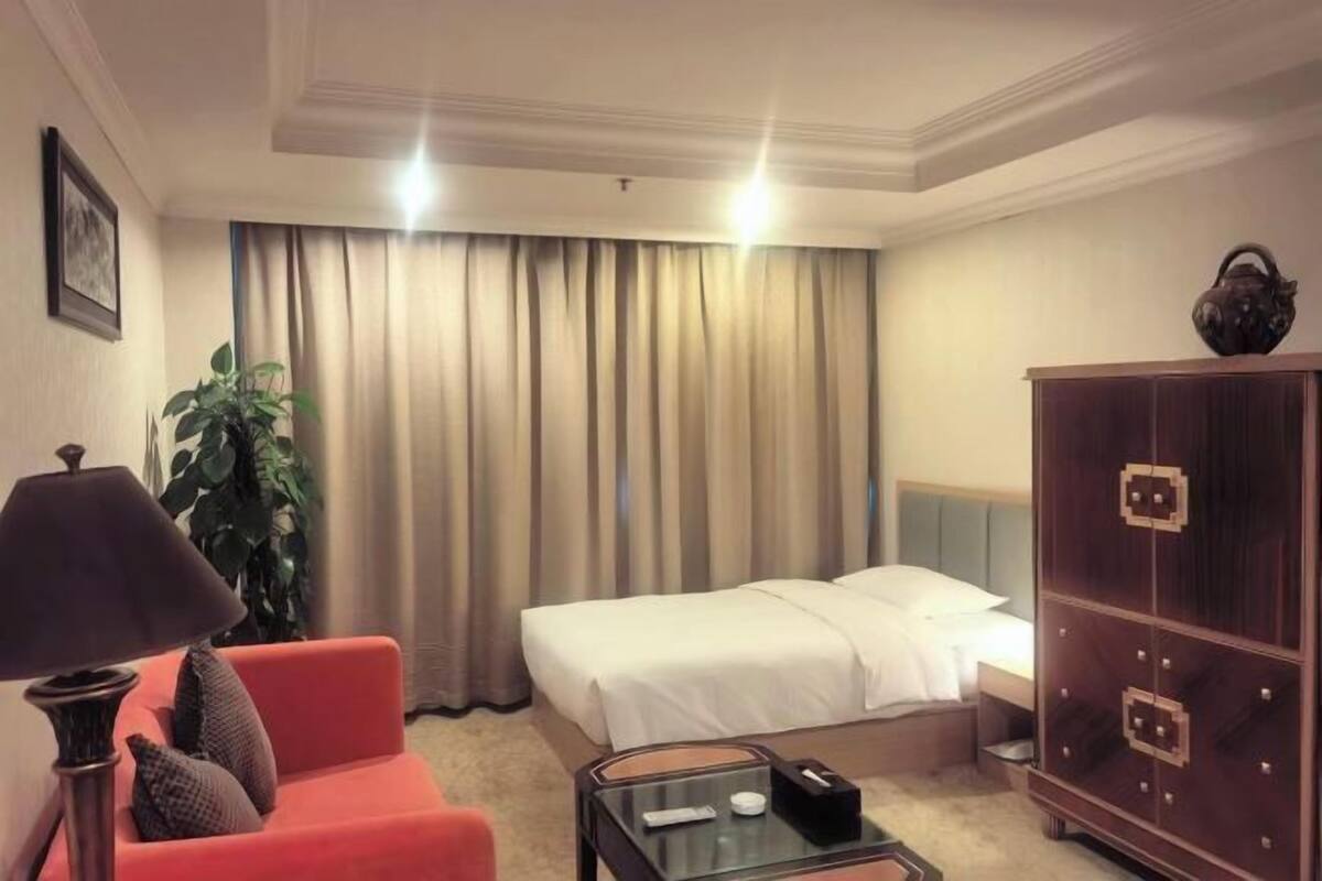Una habitación de hotel con una cama, un sillón rojo, una mesita de vidrio, un armario y una lámpara.