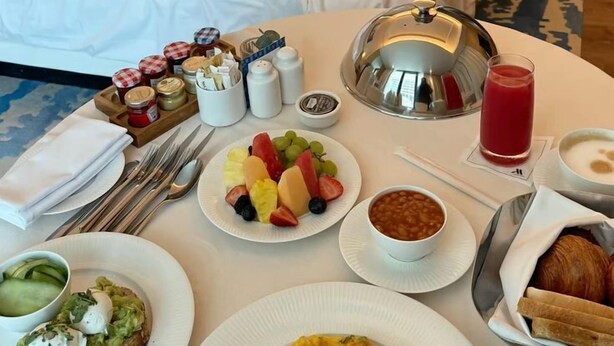 Une chambre d’hôtel avec un lit soigneusement fait, une table à manger dressée pour le petit-déjeuner, avec des œufs, des toasts et des fruits, et une cafetière.