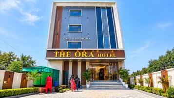A hotel entrance with a clear sky, a sign reading 'THE ORA HOTEL,' and a modern building facade.