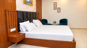 A hotel room with a wooden bed frame, a nightstand, a bedside table, and two teal chairs. - The Ora Hotel (Vrindavan)
