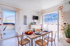 A dining area with a table set for four, a television mounted on the wall, and a view of the outdoors through large windows. - Agrilaura Apartments (Nago-Torbole)