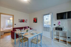 A dining area with a table set for four, a sofa, a TV mounted on the wall, and a sliding glass door. - Agrilaura Apartments (Nago-Torbole)