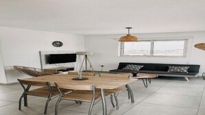 Un salon moderne avec une table de salle à manger ronde en bois, des chaises en métal, un canapé noir agrémenté de coussins à motifs, un téléviseur fixé au mur et un luminaire suspendu.
