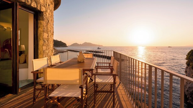 Un pontile in legno con tavolo e sedie, affacciato su uno specchio d'acqua con il tramonto sullo sfondo.