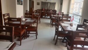 Un espace repas comprenant plusieurs tables et chaises en bois, un sol carrelé et de grandes fenêtres donnant sur les bâtiments environnants.