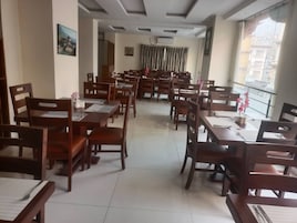 Un área de comedor con varias mesas y sillas de madera, suelo de baldosas y ventanas grandes con vistas a edificios.