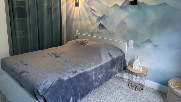 Une chambre à coucher avec un lit, une lampe fixée au mur, une petite table ronde et un tableau représentant des montagnes.