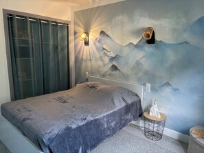 A bedroom with a bed, a wall-mounted light, a small round table, and a wall painting of mountains. - The olicleon cocoon (Aston)