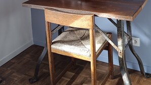 A wooden desk with a woven chair, a white wall, and a wooden beam structure.