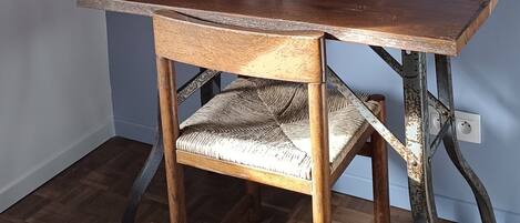 A wooden desk with a woven chair, a white wall, and a wooden beam structure.