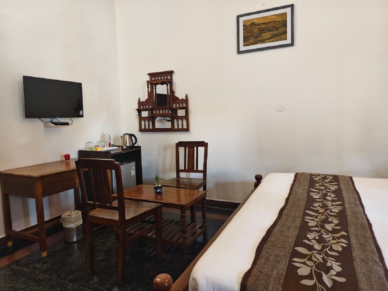 Quarto de hotel com cama, escrivaninha, cadeira, televisão, um quadro na parede e uma pequena lixeira.