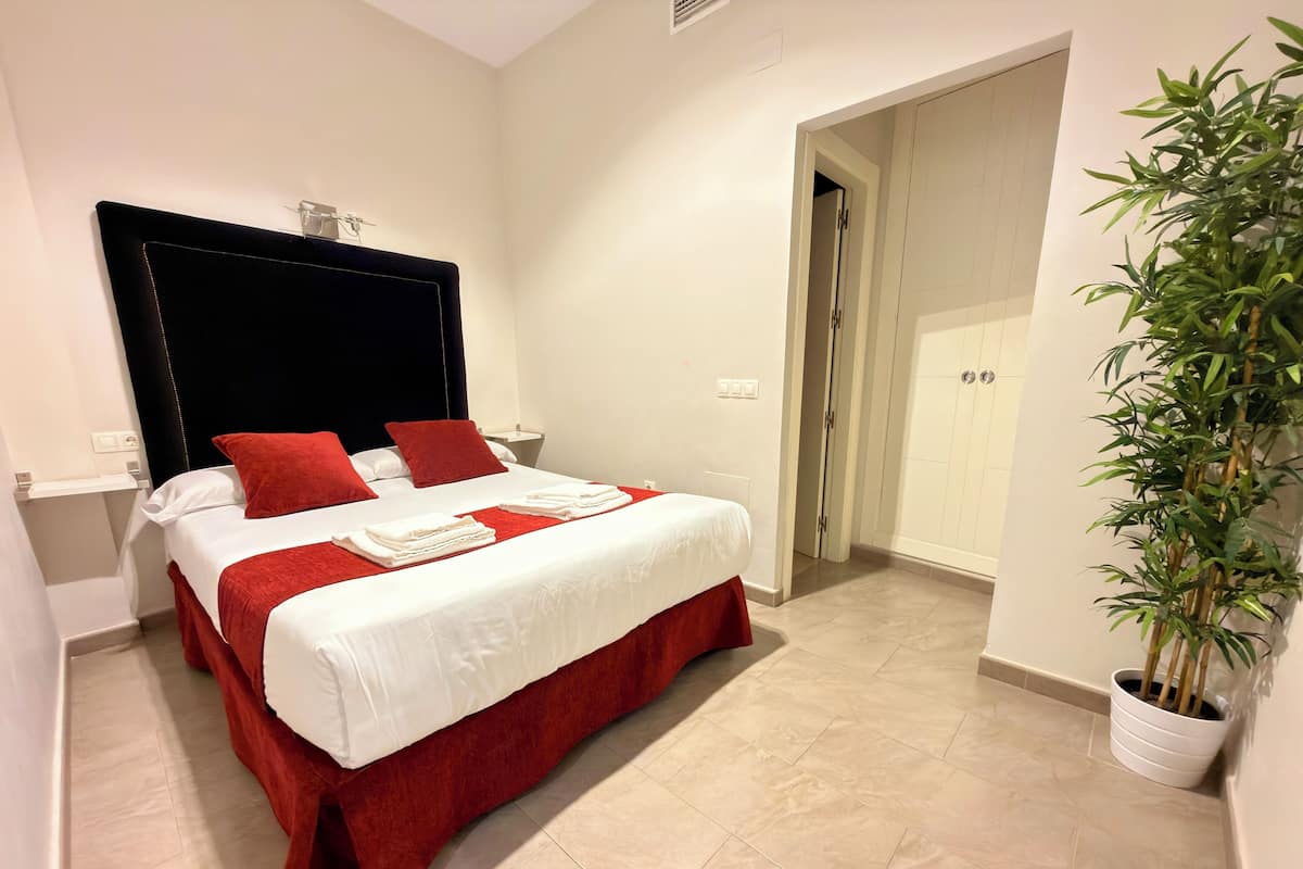 Une chambre d’hôtel avec un lit, une literie rouge et blanche, une table de chevet, une plante en pot et une porte donnant sur une autre pièce.