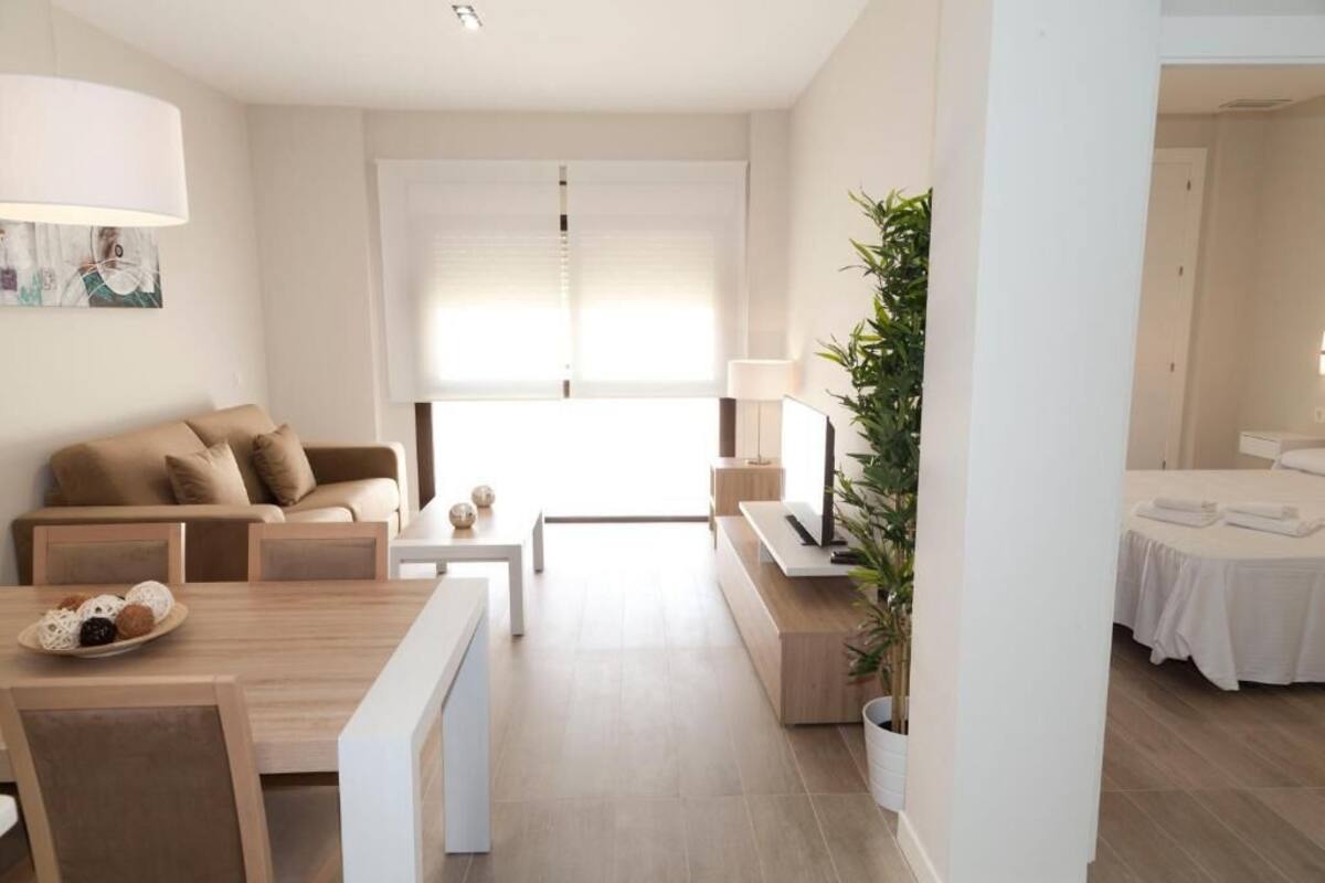 Un salon moderne et minimaliste, avec un canapé, une table basse et un coin repas. La pièce adjacente contient une plante, une lampe et un lit.
