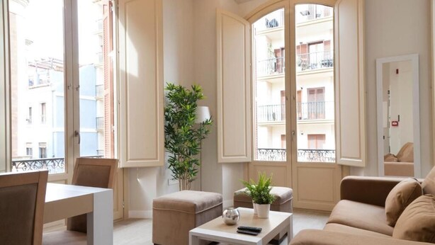 A modern living room with a sofa, ottoman, and a small table with a plant and a remote control. - Apartamentos Pinar Málaga Centro (Málaga)